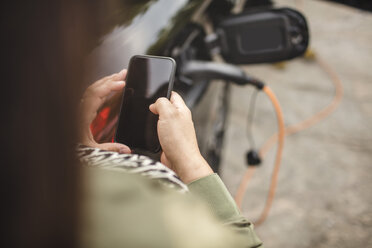 Mittelteil einer Frau, die ein Smartphone benutzt, während sie neben einem aufladenden Elektroauto steht - MASF11425