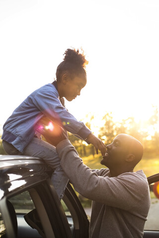 Lächelnder Mann, der einem Mädchen beim Absteigen vom Autodach hilft, bei klarem Himmel und Sonnenuntergang im Park, lizenzfreies Stockfoto