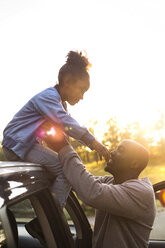 Smiling man assisting girl in getting down from car roof against clear sky during sunset at park - MASF11428