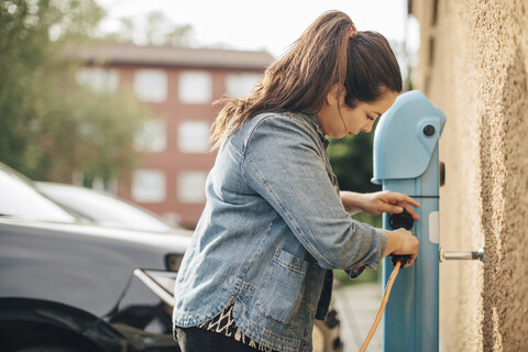 Seitenansicht einer Frau, die den Stecker für das Aufladen eines Autos an einer Station einstellt, lizenzfreies Stockfoto