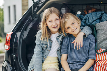 Porträt eines lächelnden blonden Mädchens, das mit dem Arm um seine Schwester im Kofferraum eines Autos vor einem Haus sitzt - MASF11472