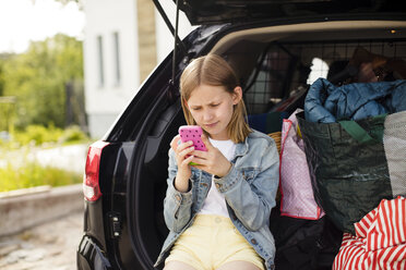 Blondes Mädchen benutzt ihr Smartphone, während sie im Kofferraum eines Elektroautos sitzt - MASF11475