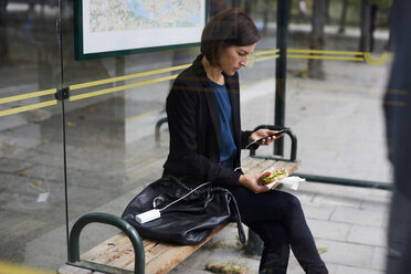 Mittlere erwachsene Geschäftsfrau, die ein Smartphone benutzt, während sie ein Sandwich an einer Bushaltestelle in der Stadt isst - MASF11478