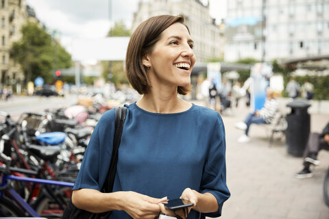 Lächelnde Geschäftsfrau im mittleren Erwachsenenalter, die wegschaut, während sie ein Smartphone in der Stadt hält, lizenzfreies Stockfoto
