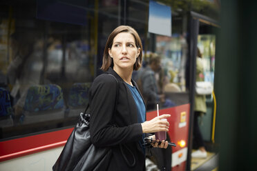 Selbstbewusste Geschäftsfrau schaut weg, während sie mit einem Getränk vor einem Bus in der Stadt steht - MASF11494