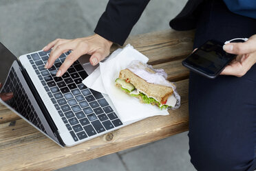 Mittelteil einer Geschäftsfrau, die einen Laptop benutzt, während sie mit Smartphone und Sandwich an einer Bushaltestelle in der Stadt sitzt - MASF11495