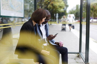 Multiethnische Pendler teilen sich ein Smartphone, während sie an einer Bushaltestelle in der Stadt sitzen - MASF11496