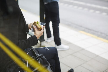 Mittelteil einer Geschäftsfrau, die ein Sandwich isst, während sie mit ihrem Smartphone an einer Bushaltestelle in der Stadt sitzt - MASF11497
