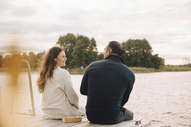 Lächelndes Paar im Gespräch auf dem Steg am See gegen den Himmel sitzend - MASF11501