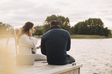 Rückansicht eines Paares, das Musik auf einem Smartphone hört, während es auf einem Steg am See sitzt, gegen den Himmel - MASF11502