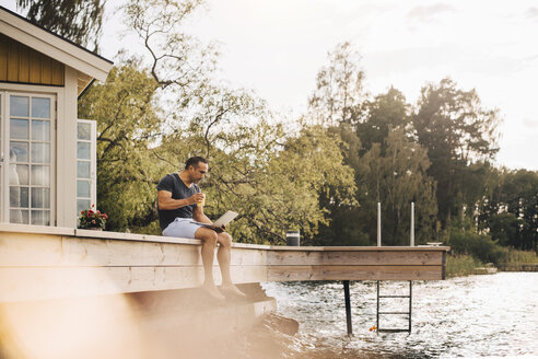 Mature man having juice while using laptop on patio by lake - MASF11504