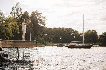 Full length of woman talking through video call on mobile phone while standing at patio by lake against sky - MASF11507