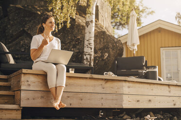 Full length of woman talking on headphones while using laptop at holiday villa - MASF11508