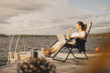 Full length of woman with laptop talking through headphones while sitting on jetty against sky - MASF11511