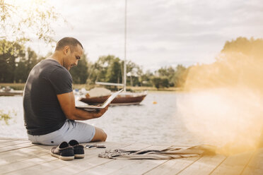 Reifer Mann, der einen Laptop benutzt, während er im Urlaub auf einem Steg über einem See sitzt - MASF11516