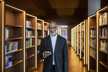 Porträt eines lächelnden Geschäftsmannes mit Smartphone in einer Bibliothek - FMKF05351