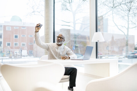 Porträt eines reifen Mannes mit Laptop, der am Schreibtisch sitzt und ein Selfie mit seinem Smartphone macht, lizenzfreies Stockfoto