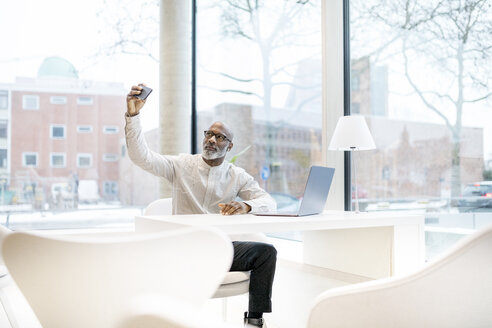 Porträt eines reifen Mannes mit Laptop, der am Schreibtisch sitzt und ein Selfie mit seinem Smartphone macht - FMKF05361