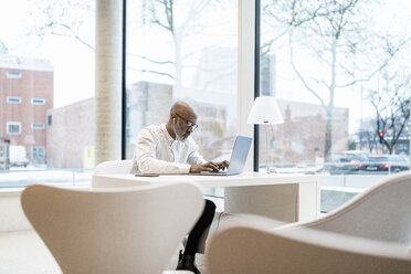 Älterer Geschäftsmann mit Kopfhörern sitzt am Schreibtisch und arbeitet am Laptop - FMKF05362