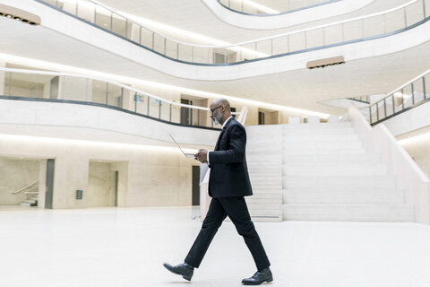 Intelligenter reifer Geschäftsmann, der einen Laptop benutzt, während er durch das Foyer geht, lizenzfreies Stockfoto