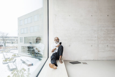 Barefoot mature businessman sitting on window sill using smartphone - FMKF05373