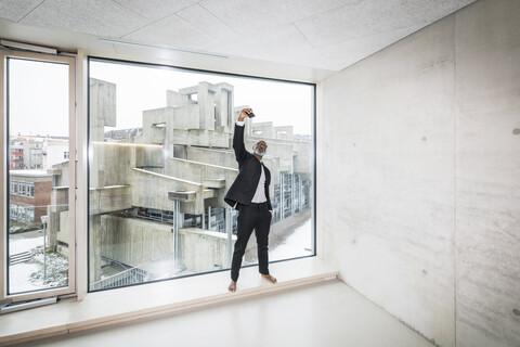 Barfüßiger Geschäftsmann steht vor einem Fenster und macht ein Selfie mit seinem Smartphone, lizenzfreies Stockfoto