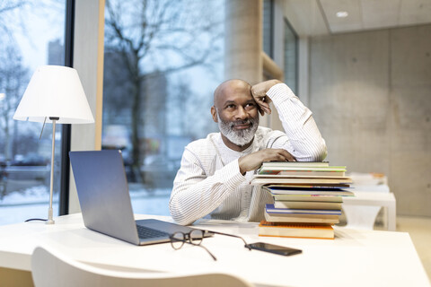 Portrait of smiling mature man sitting at desk with laptop and stack of books stock photo