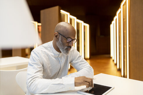 Älterer Mann sitzt am Schreibtisch in einer Bibliothek und benutzt ein digitales Tablet - FMKF05379