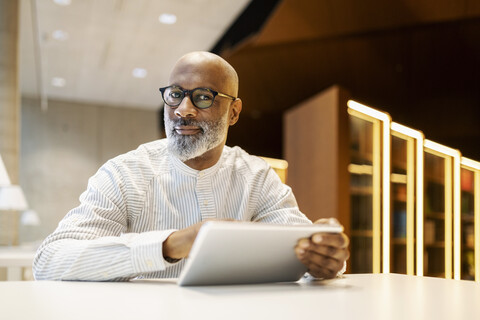 Portrait of mature man sitting at desk in a library with digital tablet stock photo