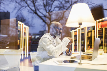 Älterer Mann sitzt abends am Schreibtisch in einer Bibliothek und benutzt einen Laptop - FMKF05384