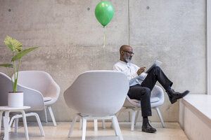 Älterer Geschäftsmann mit grünem Ballon, der auf einem Sessel sitzt und ein digitales Tablet benutzt - FMKF05392