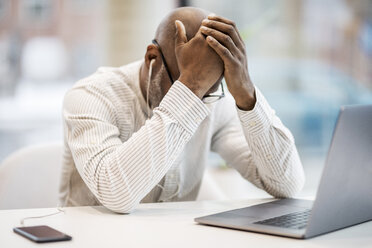 Mature businessman with earphones, smartphoen and laptop sitting with hands on head at desk - FMKF05396