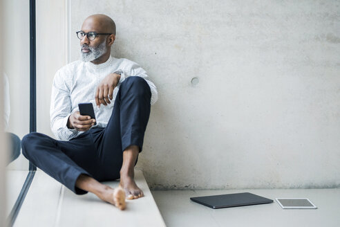 Barefoot mature businessman with smartphone sitting on window sill looking at distance - FMKF05403