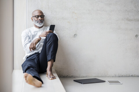 Barefoot mature businessman sitting on window sill looking at smartphone stock photo