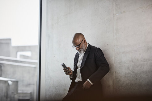 Bald mature businessman leaning against concrete wall looking at cell phone - FMKF05405