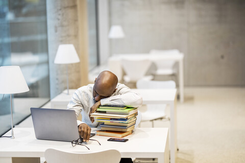 Erschöpfter reifer Mann sitzt am Schreibtisch und stützt sich auf einen Bücherstapel, lizenzfreies Stockfoto