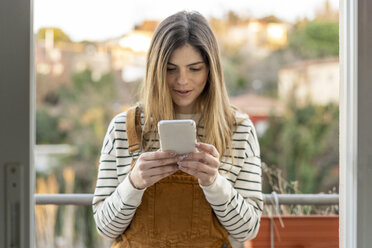 Porträt einer jungen Frau, die auf einem Balkon steht und auf ihr Mobiltelefon schaut - AFVF02394