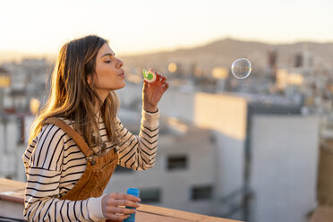 Young woman blowing soap bubbles on roof terrace in the evening - AFVF02404