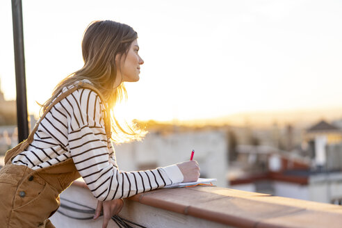 Junge Frau macht sich auf der Dachterrasse bei Sonnenuntergang Notizen - AFVF02406