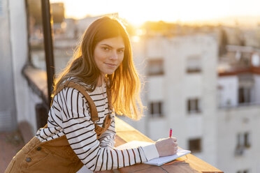 Porträt einer jungen Frau, die auf einer Dachterrasse bei Sonnenuntergang Notizen macht - AFVF02409