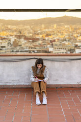 Young woman sitting on roof terrace taking notes - AFVF02410