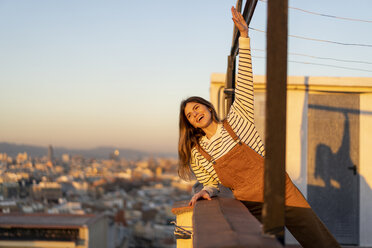 Porträt einer lachenden jungen Frau, die sich bei Sonnenuntergang auf einer Dachterrasse entspannt - AFVF02413