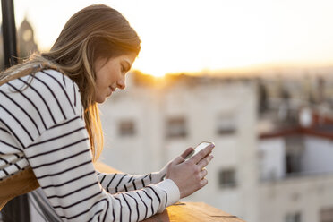 Junge Frau auf dem Balkon bei Sonnenuntergang mit Mobiltelefon - AFVF02420
