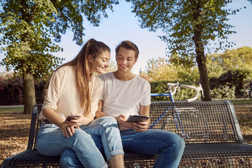 Junges Paar im Park auf einer Bank sitzend mit Handy und Tablet - JHAF00041