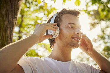 Lächelnder junger Mann mit Kopfhörern, der draußen Musik hört - JHAF00043
