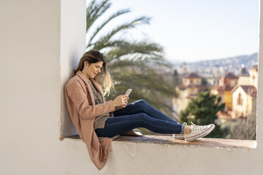 Laughing young woman sitting on a wall using cell phone - AFVF02428