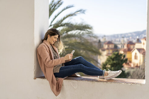 Lachende junge Frau, die auf einer Mauer sitzt und ein Mobiltelefon benutzt - AFVF02428