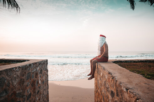 Thailand, als Weihnachtsmann verkleideter Mann, der auf einer Mauer vor dem Meer sitzt und den Sonnenuntergang beobachtet - HMEF00209