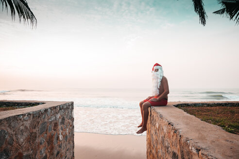 Thailand, als Weihnachtsmann verkleideter Mann, der bei Sonnenuntergang auf einer Mauer vor dem Meer sitzt - HMEF00210