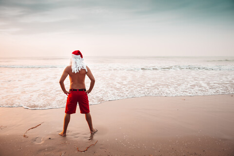 Thailand, Rückenansicht eines als Weihnachtsmann verkleideten Mannes, der bei Sonnenuntergang am Strand steht und auf den Horizont schaut, lizenzfreies Stockfoto